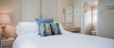 A hotel room with a large bed, a bedside table with a lamp, a small bedside table with a tray, a mirror, and a door leading to a bathroom.