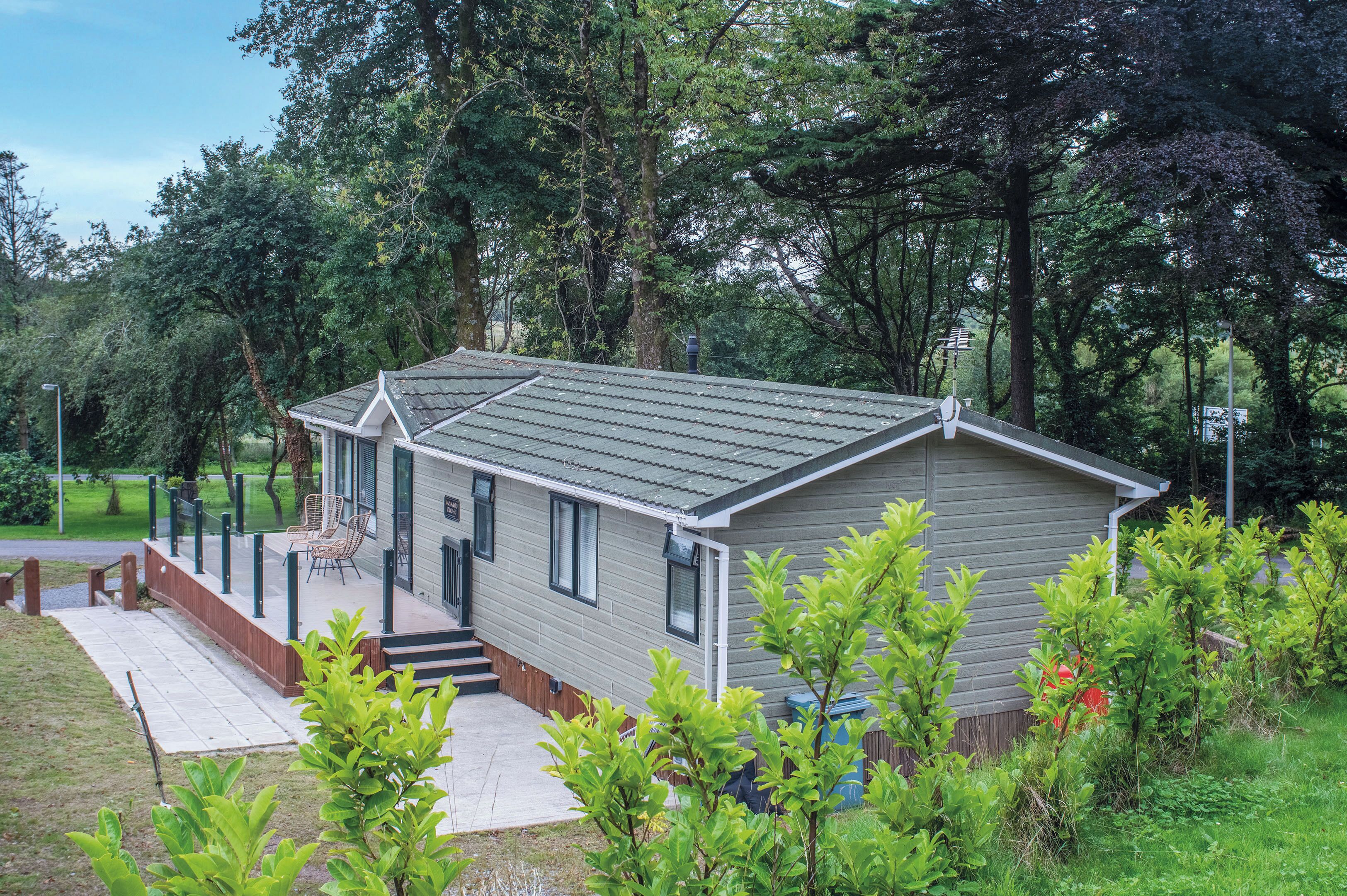 A single-story mobile home with a porch, a small deck, and a front yard with young trees.