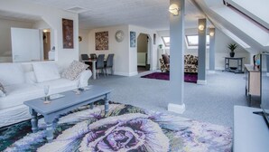 A spacious living area with a white sofa, a coffee table, and a colorful abstract rug. There is a dining area with chairs and a table, and a view of the city through large windows. - The Penthouse - 1 Bedroom Penthouse - Milford Haven (Milford Haven)