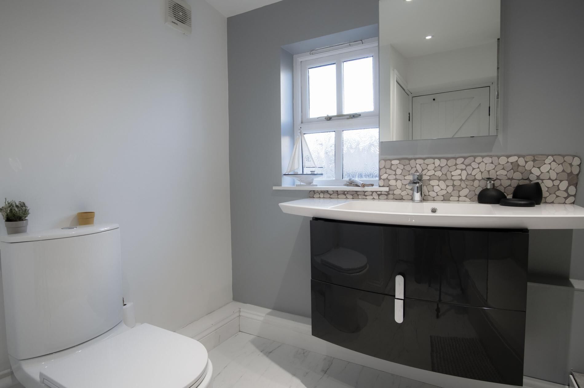 Un baño moderno con inodoro blanco, lavamanos blanco y un salpicadero de piedra, y una ventana sobre el lavamanos.