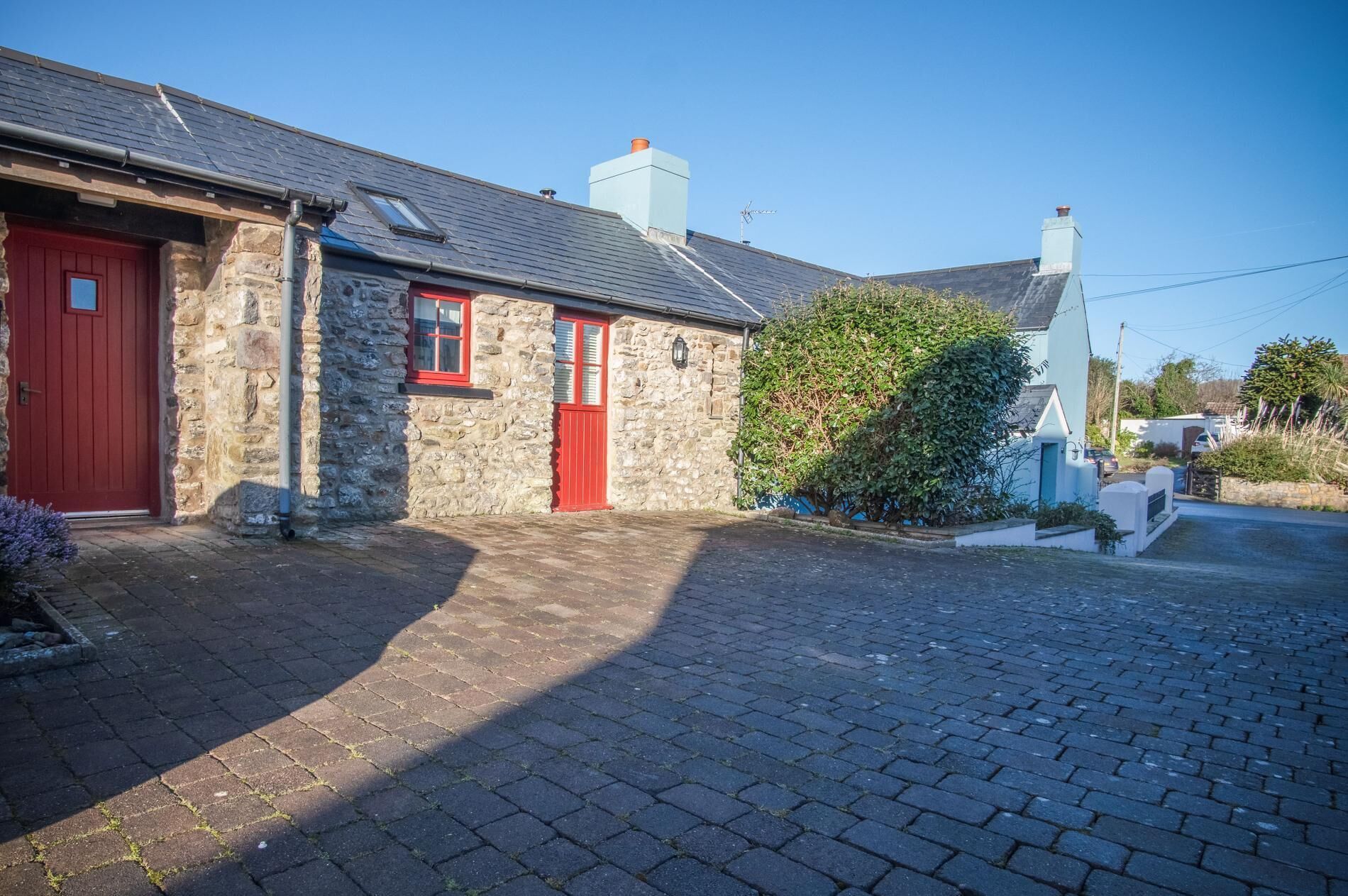 A stone building with a red door and windows, a paved driveway, and a hedge.