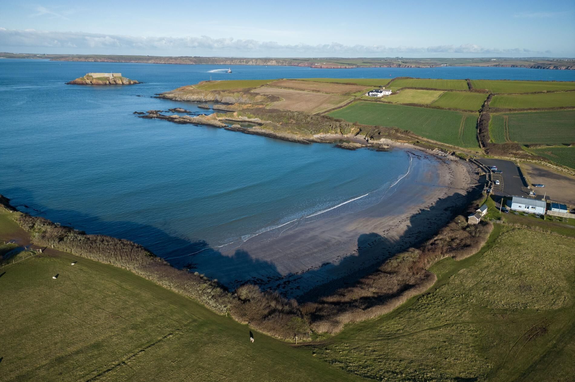 Vue aérienne d’une zone côtière avec une plage de sable, des champs couverts d’herbe et un petit bâtiment blanc situé près du rivage.