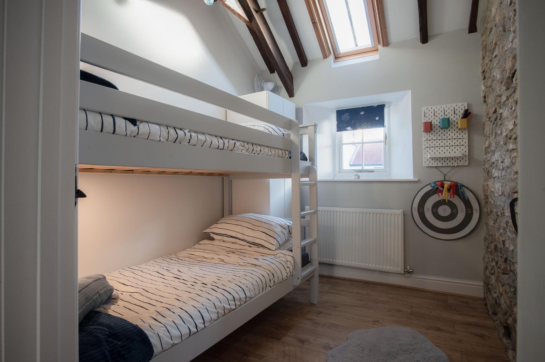 Une chambre avec un lit superposé, une fenêtre, un radiateur et un jeu de fléchettes accroché au mur.