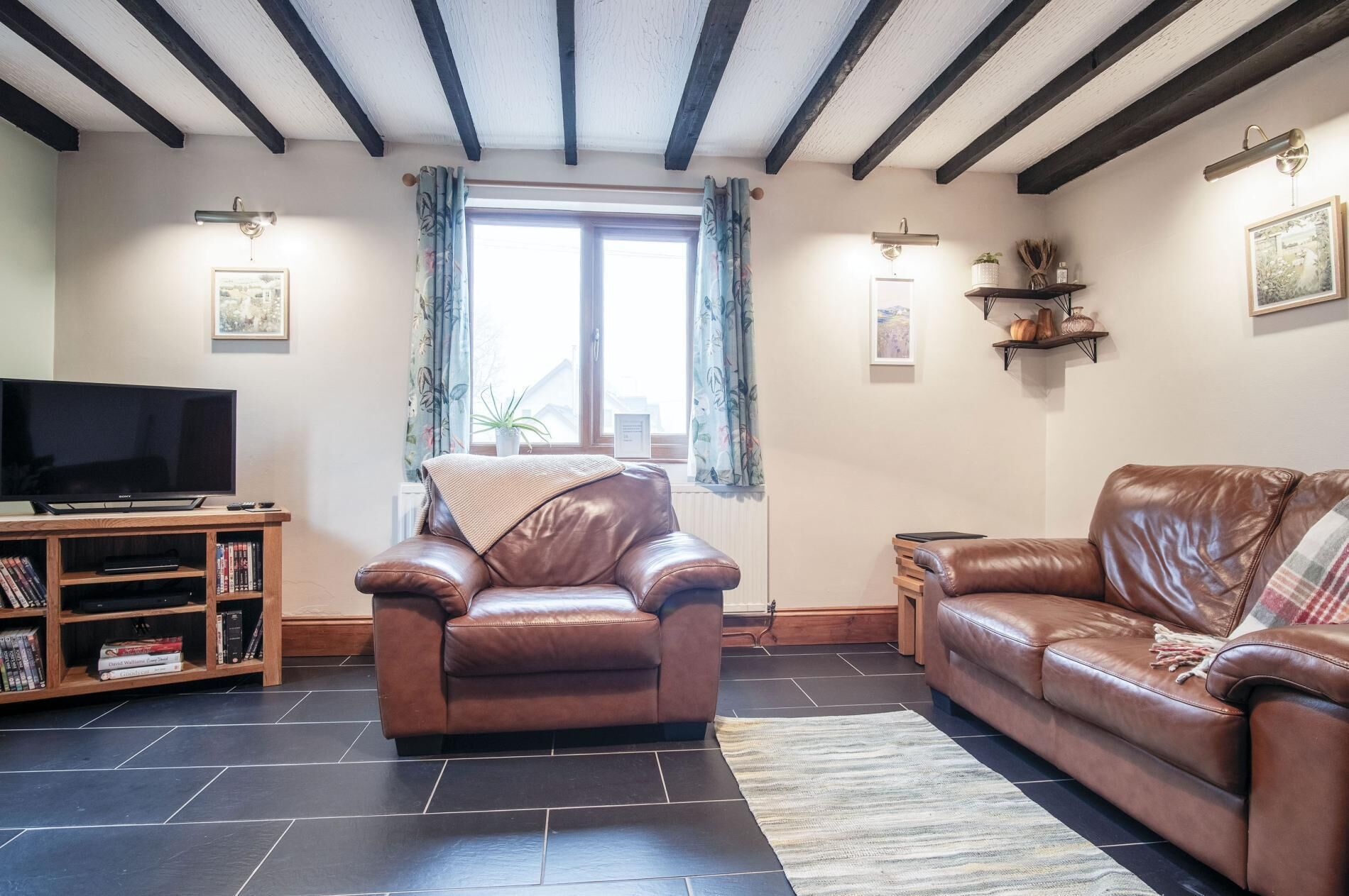 A living room with a leather sofa, a leather armchair, a TV on a wooden stand, and a bookshelf.