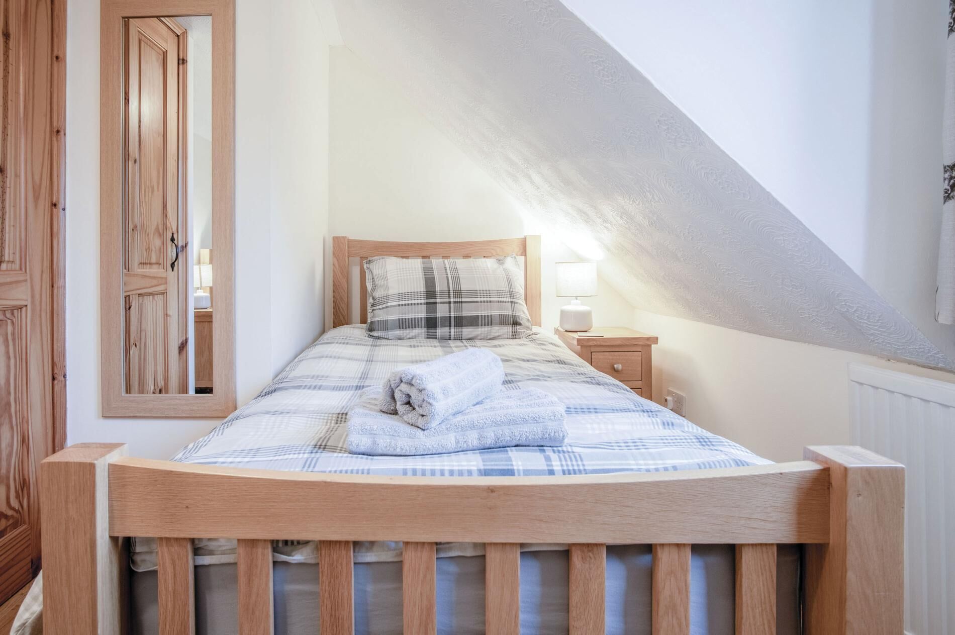 A bedroom with a wooden bed frame, a plaid bedspread, a white pillow, and a bedside lamp.