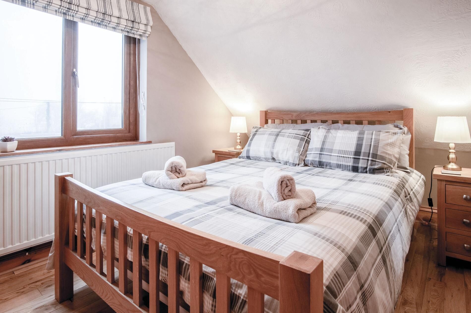 A cozy bedroom with a wooden bed, plaid bedding, two rolled towels, a nightstand with a lamp, and a window with curtains.