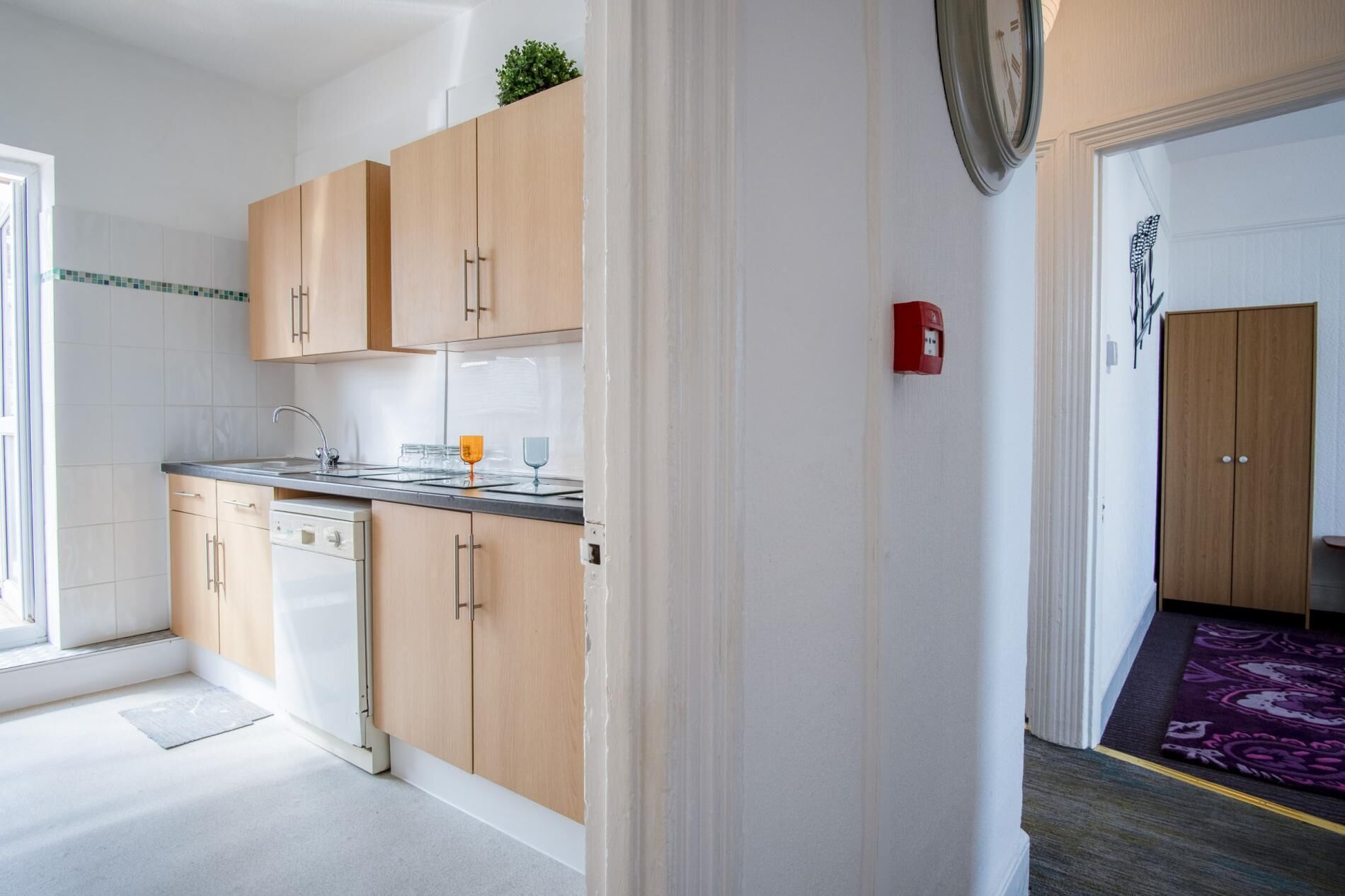 A modern kitchen with light wood cabinets, a stainless steel sink, and a red fire alarm.