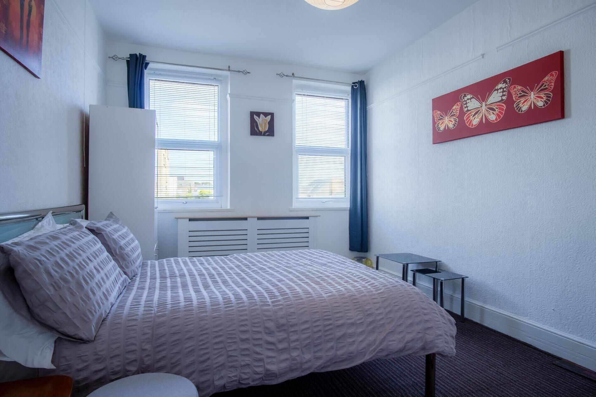 Un dormitorio con cama, dos ventanas con persianas, un radiador, una mesita y un cuadro de una mariposa roja en la pared.