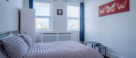 Un dormitorio con cama, dos ventanas con persianas, un radiador, una mesita y un cuadro de una mariposa roja en la pared.