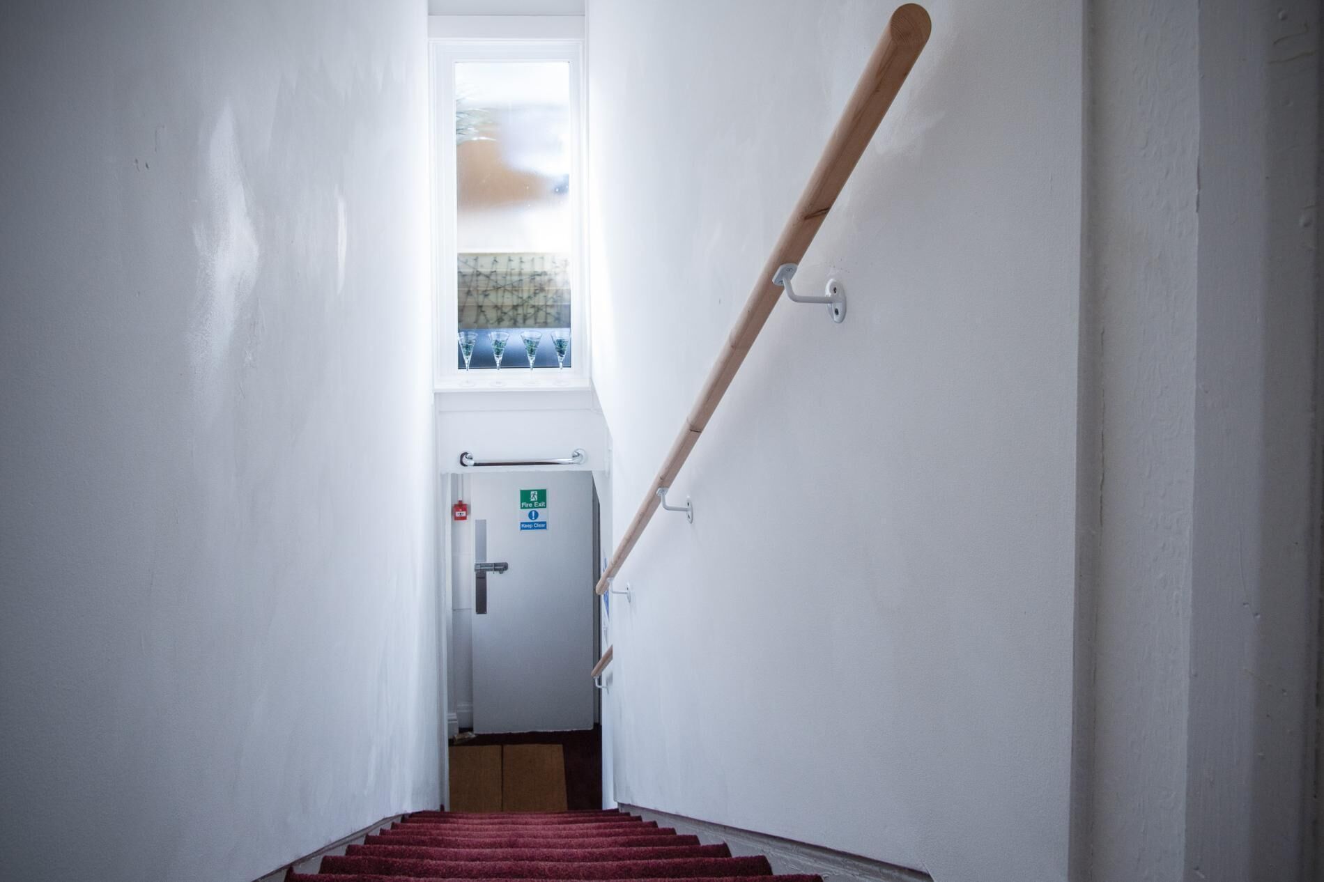 A staircase with a wooden handrail, a fire exit sign, and a window at the top.
