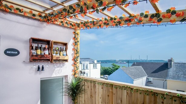 A balcony with a wooden pergola, a wine bottle shelf, and a view of the sea and buildings. - Skyfall - 2 Bedroom Apartment - Milford Haven (Milford Haven)