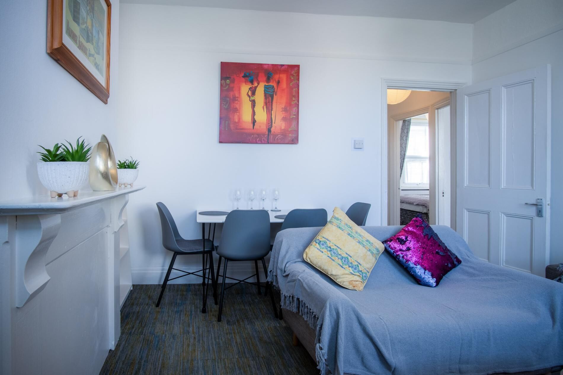 A compact hotel room with a bed, a desk with chairs, a small table with potted plants, and a painting on the wall.