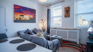 A modern living room with a sofa, a dining table with round black placemats, a large painting of a sunset, and a radiator. - Skyfall - 2 Bedroom Apartment - Milford Haven (Milford Haven)