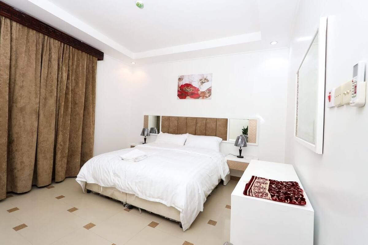 Una habitación de hotel moderna con una cama grande, una mesita de noche, un espejo y un termostato fijado a la pared.