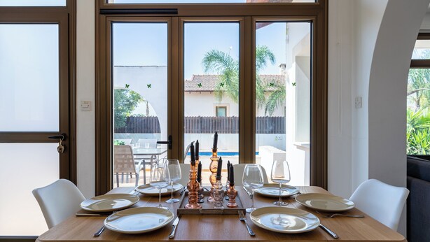 Une table à manger dressée avec des assiettes, des couverts et des bougies, donnant sur un jardin à travers de grandes fenêtres.