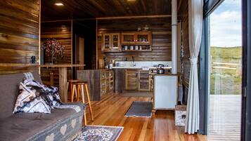 A cozy cabin interior with a wooden interior, a sofa with patterned cushions, a kitchen area with wooden cabinets, and a view through a large window.