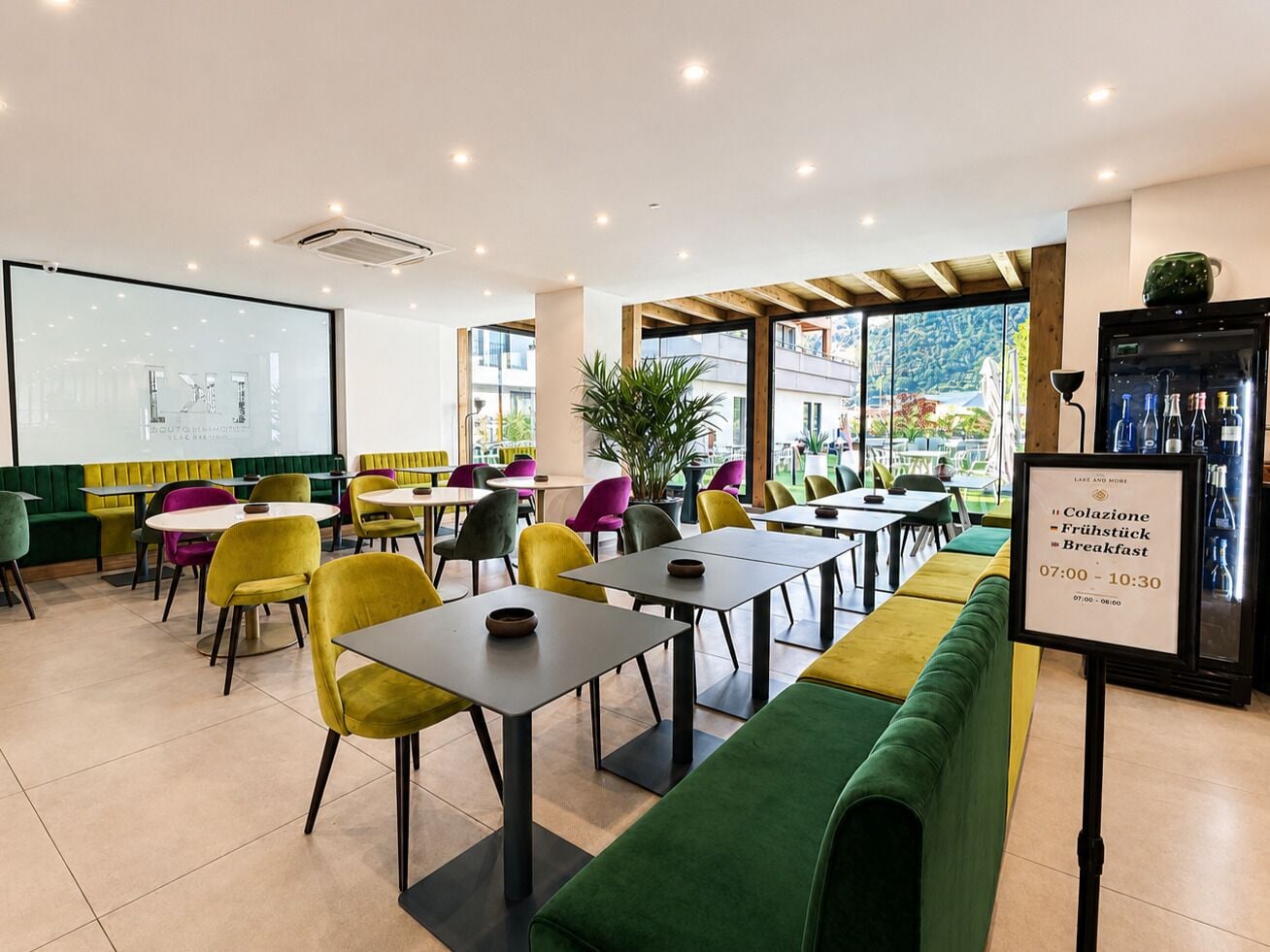 A modern dining area with multiple tables and chairs, a green sofa, and a sign indicating breakfast hours.