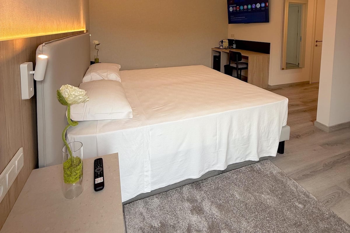 Une chambre d’hôtel moderne avec un grand lit, une table de chevet dotée d’une télécommande, un vase avec une fleur et un téléviseur à écran plat fixé au mur.