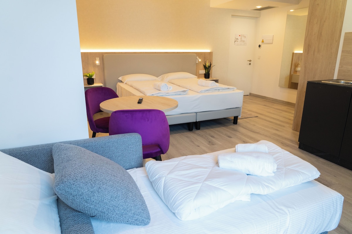 A modern hotel room with a bed, a small round table, a purple chair, and a sofa.