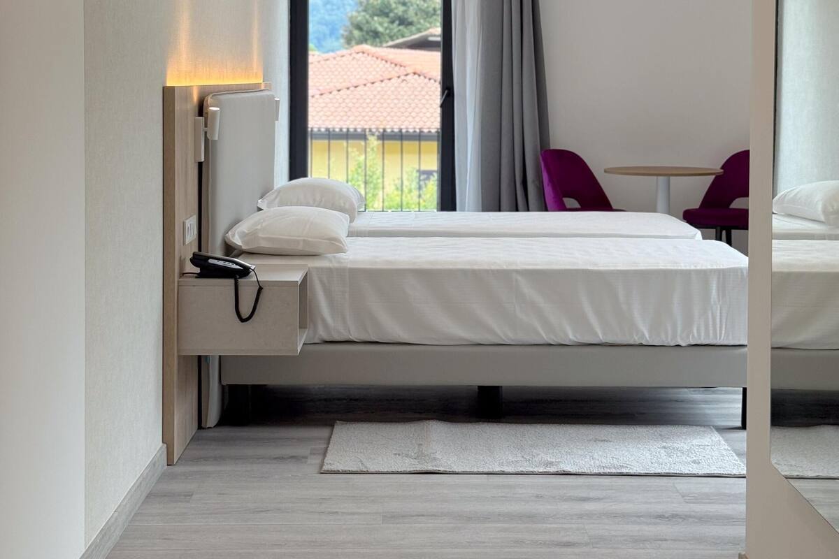 Une chambre d’hôtel moderne, équipée d’un lit, d’une table de chevet avec une lampe, offrant une vue sur l’extérieur et dotée d’une fenêtre avec des rideaux.
