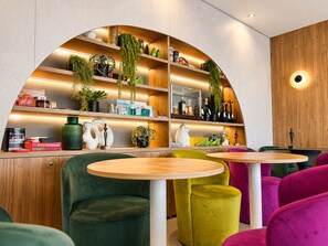 A modern interior with a curved wooden shelf displaying various items, a round wooden table, and vibrant green and pink upholstered chairs. - Lake and More Suite Hotel (Paratico)
