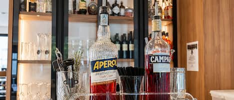A bar area with bottles of Aperol and Campari, a glass rack, and a wooden shelf with various bottles in the background.