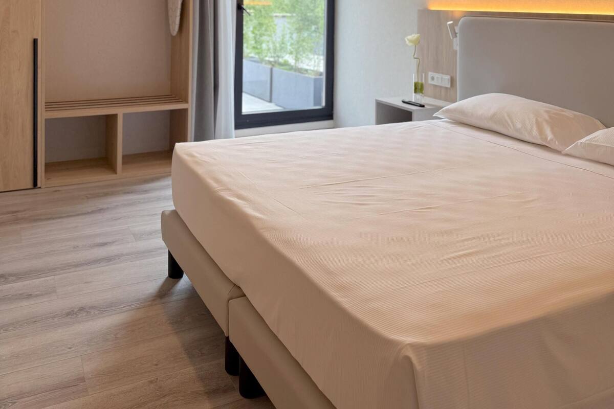 Une chambre d’hôtel moderne dotée d’un grand lit, d’une armoire intégrée, d’une table de chevet avec une lampe et offrant une vue sur la verdure grâce à une grande fenêtre.