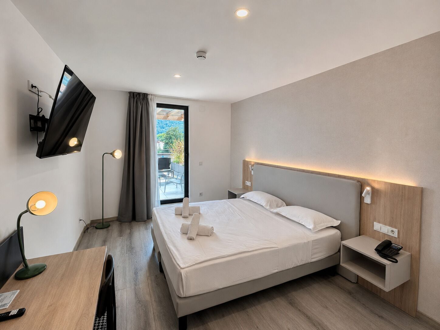 Una habitación de hotel moderna con una cama, una mesita de noche con un teléfono, un escritorio con una lámpara y un televisor montado en la pared.