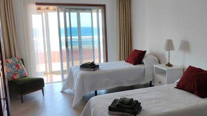Habitación de hotel con dos camas, una silla, una mesita de noche y vista al océano.
