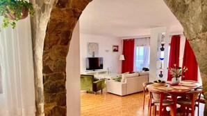 Un espacio de sala acogedor con un arco de piedra, un sofá, una mesa de comedor con sillas, un televisor y una zona de cocina al fondo.