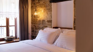 Un dormitorio con pared de piedra, cama con ropa blanca, ventana con cortinas y una lámpara colgante.