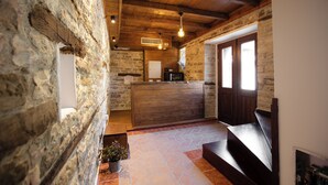 Un interior rústico con paredes de piedra, vigas de madera, un mostrador de recepción y una escalera que sube a un nivel superior.