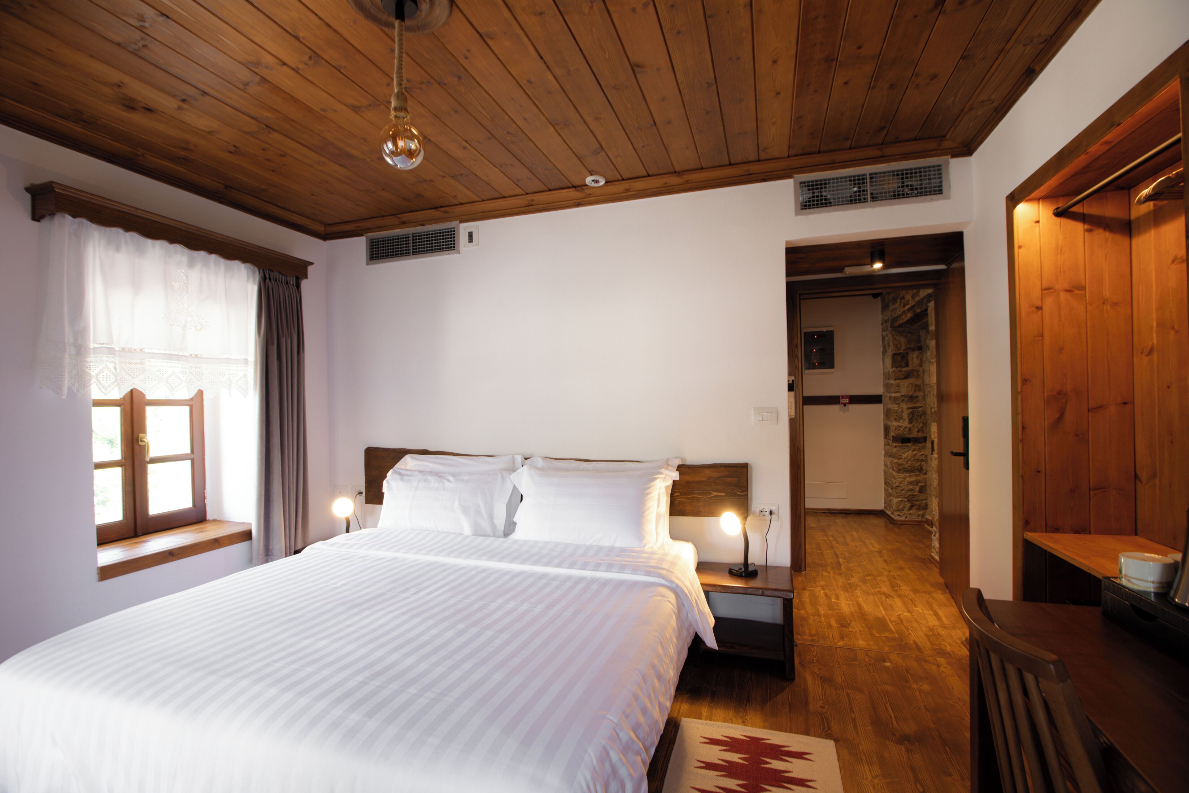 Un dormitorio con techo de madera, una cama grande, una mesita de noche con lámpara y una pared de piedra al fondo.