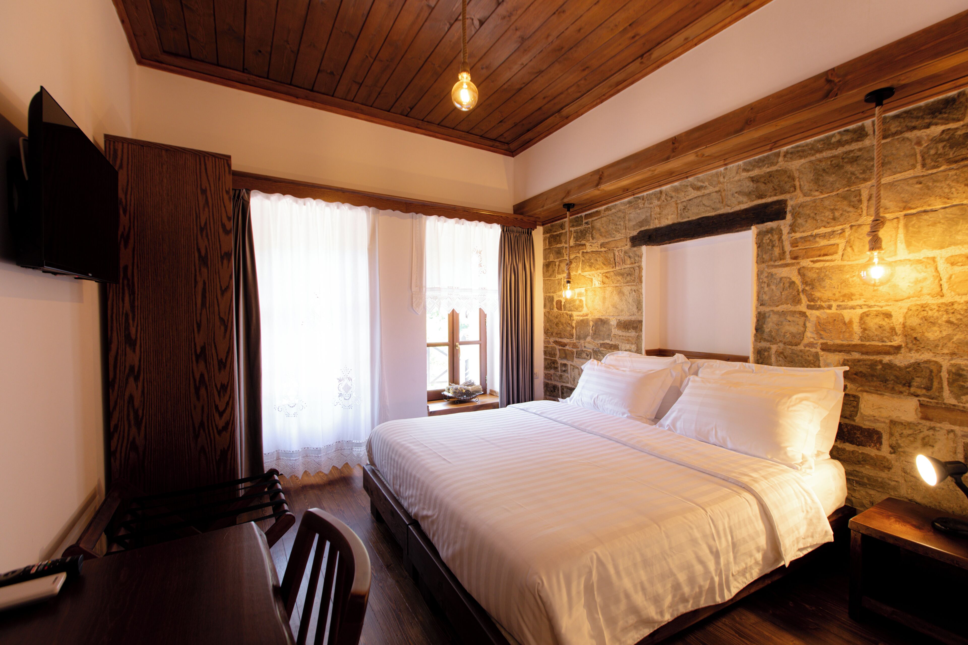 Une chambre avec un plafond en bois, un mur en pierre, un lit recouvert de draps blancs, un téléviseur fixé au mur et une fenêtre avec des rideaux.