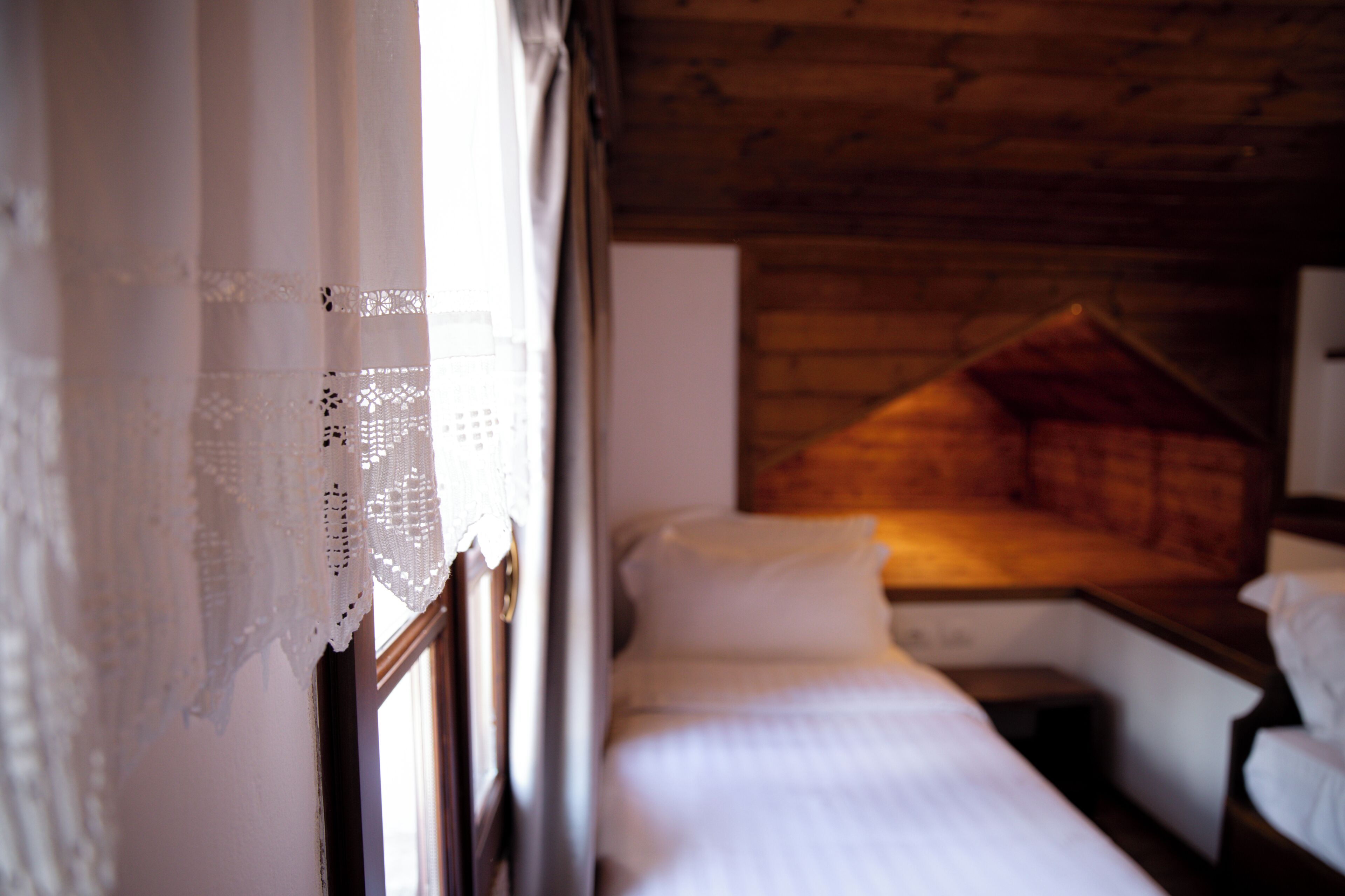 Una habitación con techo de madera, cama con ropa blanca y ventana con cortinas de encaje.