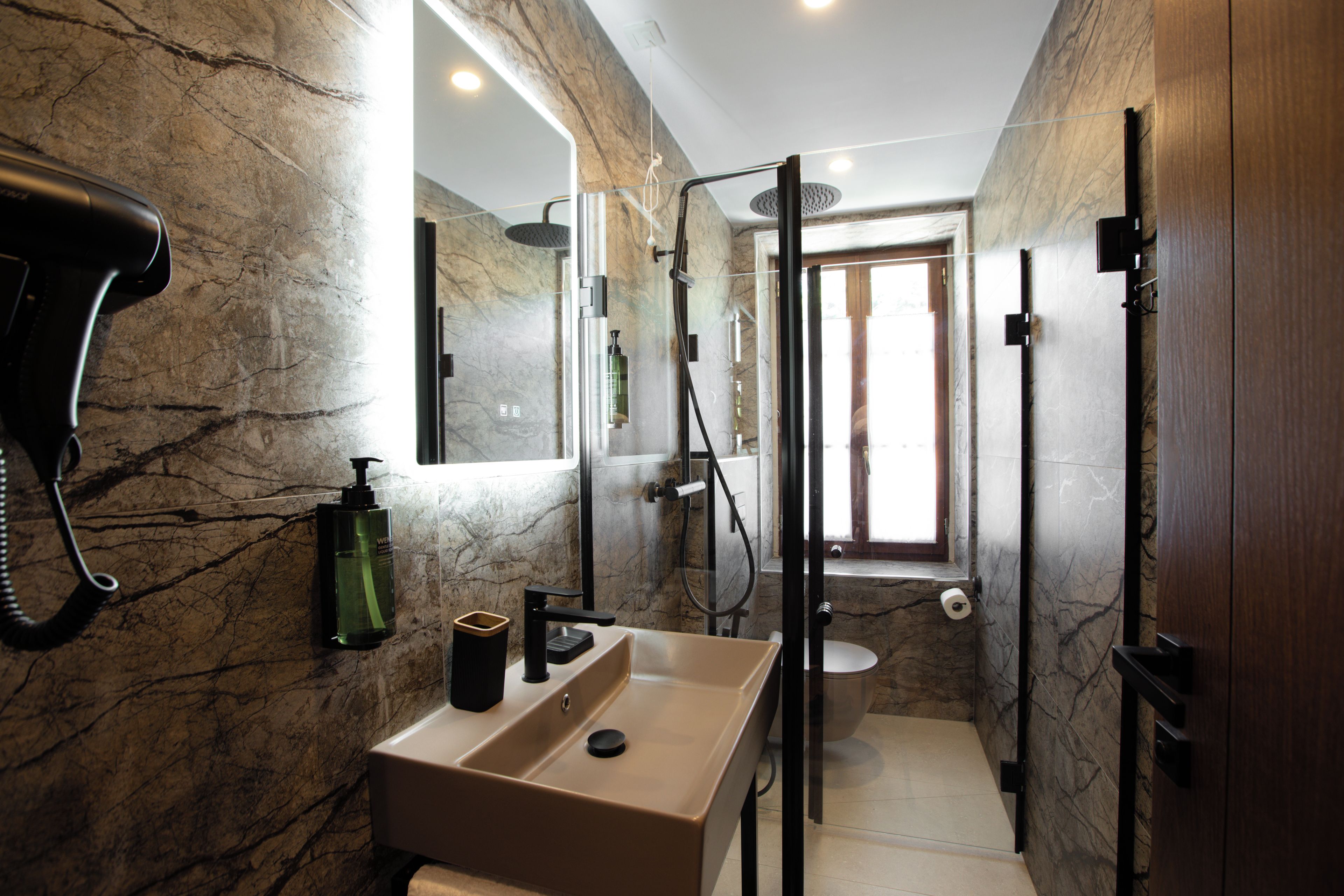 Un baño con pared de mármol, un lavamanos con espejo, un secador de pelo y una zona de ducha.