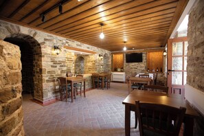 Un interior rústico con paredes de piedra, vigas de madera en el techo y un arco de piedra. Hay mesas y sillas de madera, un televisor de pantalla plana montado en la pared y una estantería para vinos con botellas.