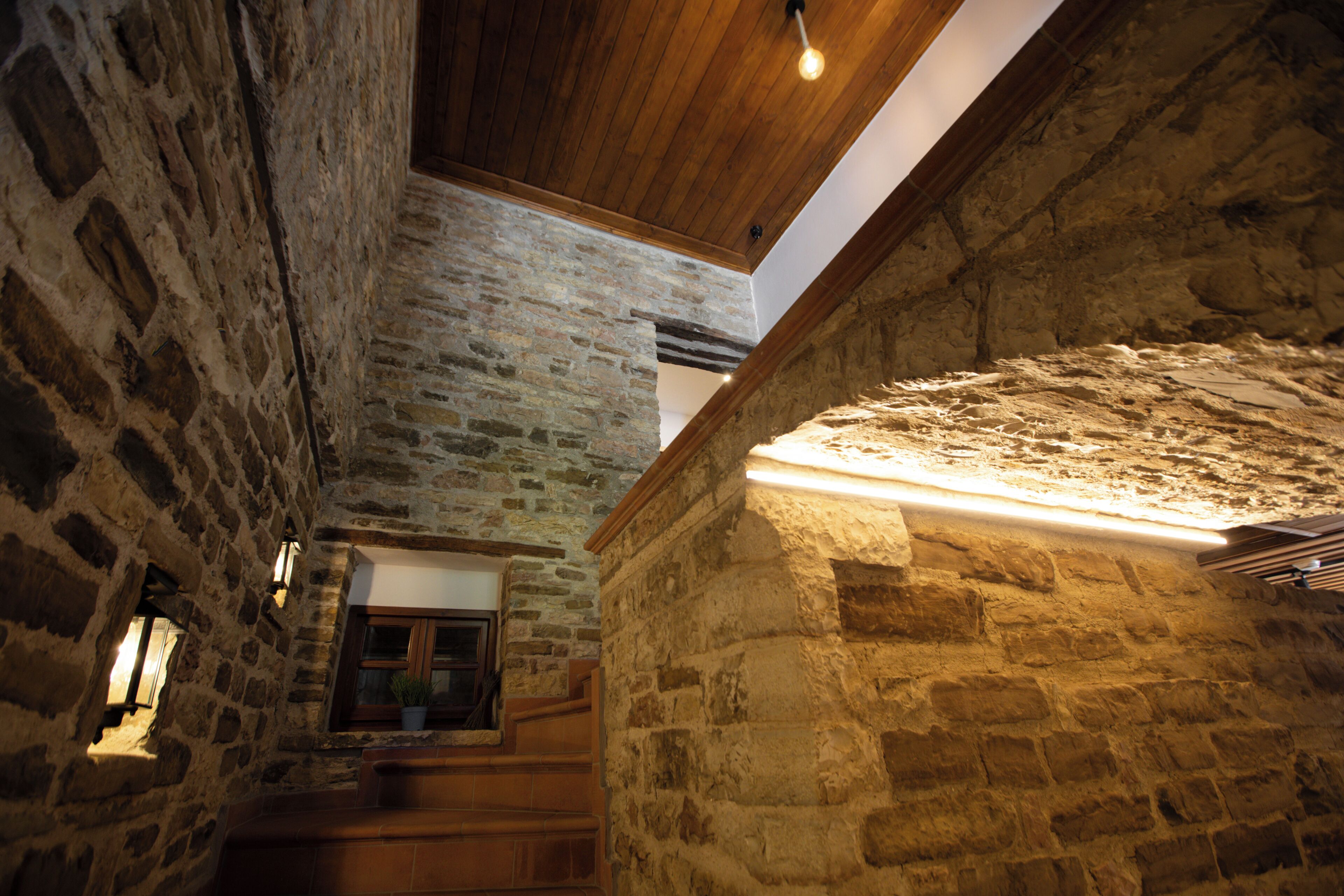 Interior de piedra con una escalera de madera, una ventana con vistas y un candelabro.