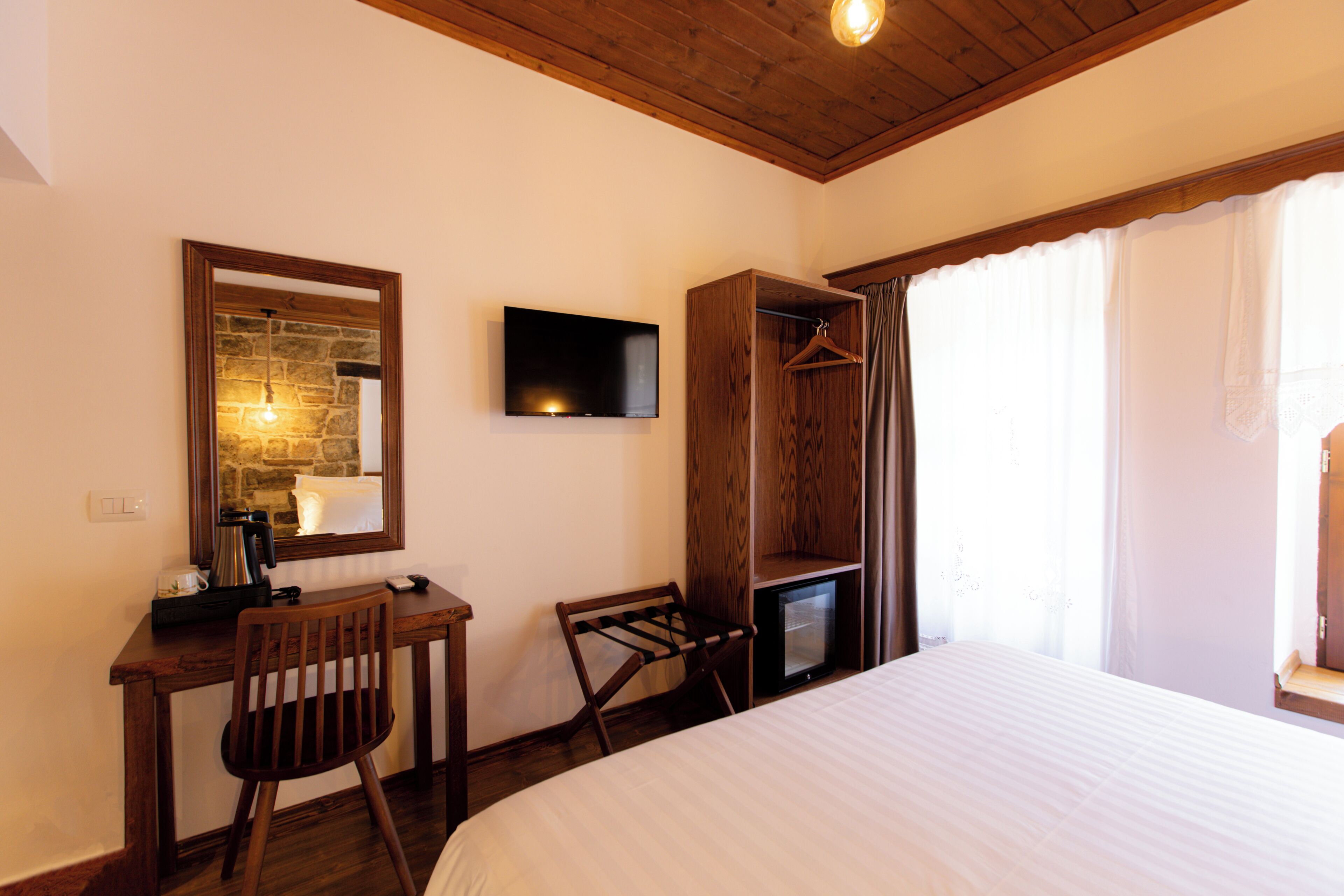 A hotel room with a bed, a desk with a chair, a television, a coat rack, and a window with curtains.