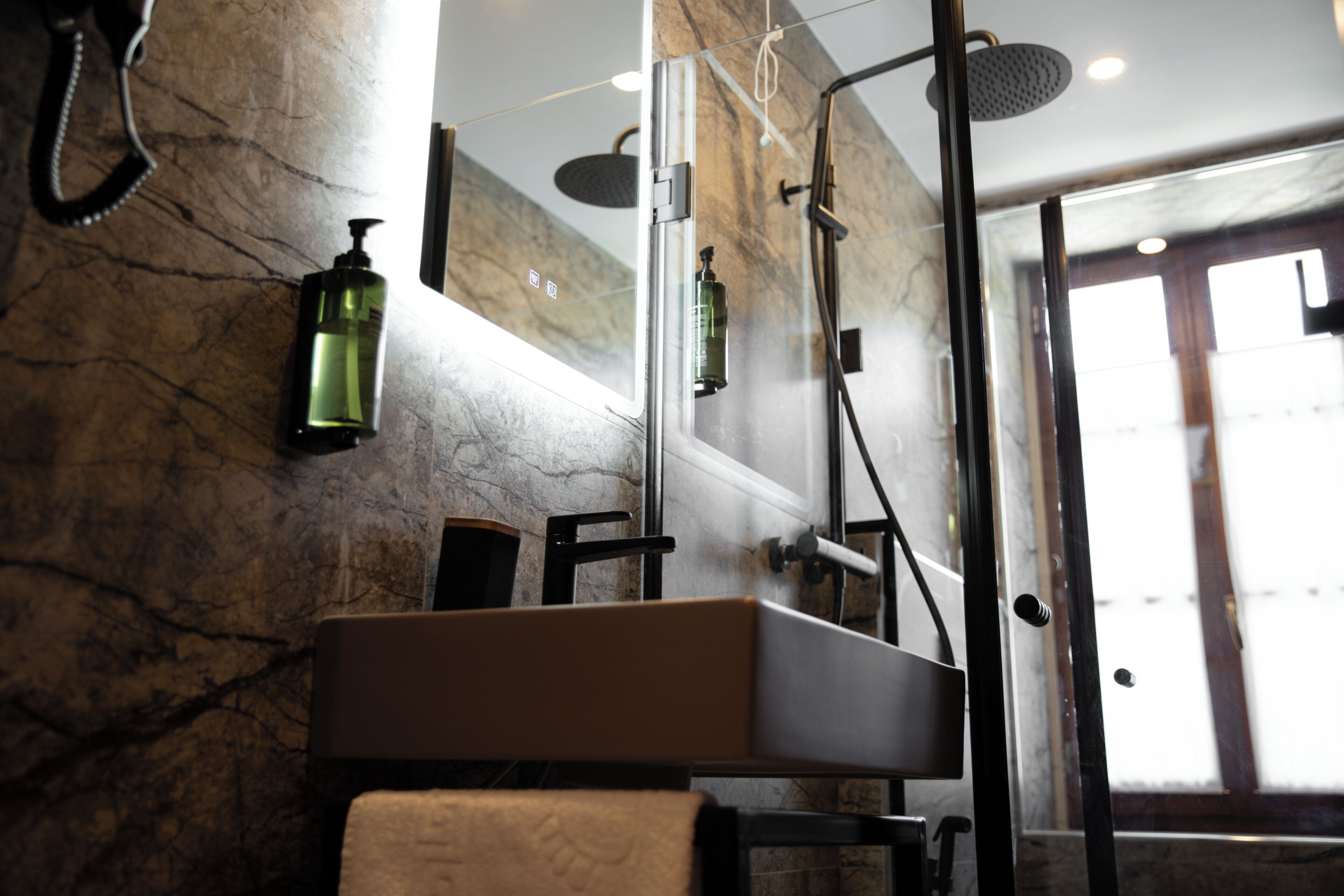 A bathroom with a marble wall, a large mirror, a shower with a handheld showerhead, and a bottle of shampoo.