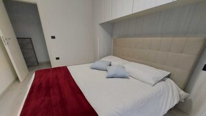 Un letto rifatto con cura, con una coperta rossa e due cuscini, una testiera bianca e una porta che dà su un corridoio.