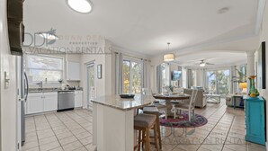A spacious kitchen with white cabinets, a dishwasher, and a large island with stools. There is a dining area with a table and chairs, and a balcony with a view of palm trees.