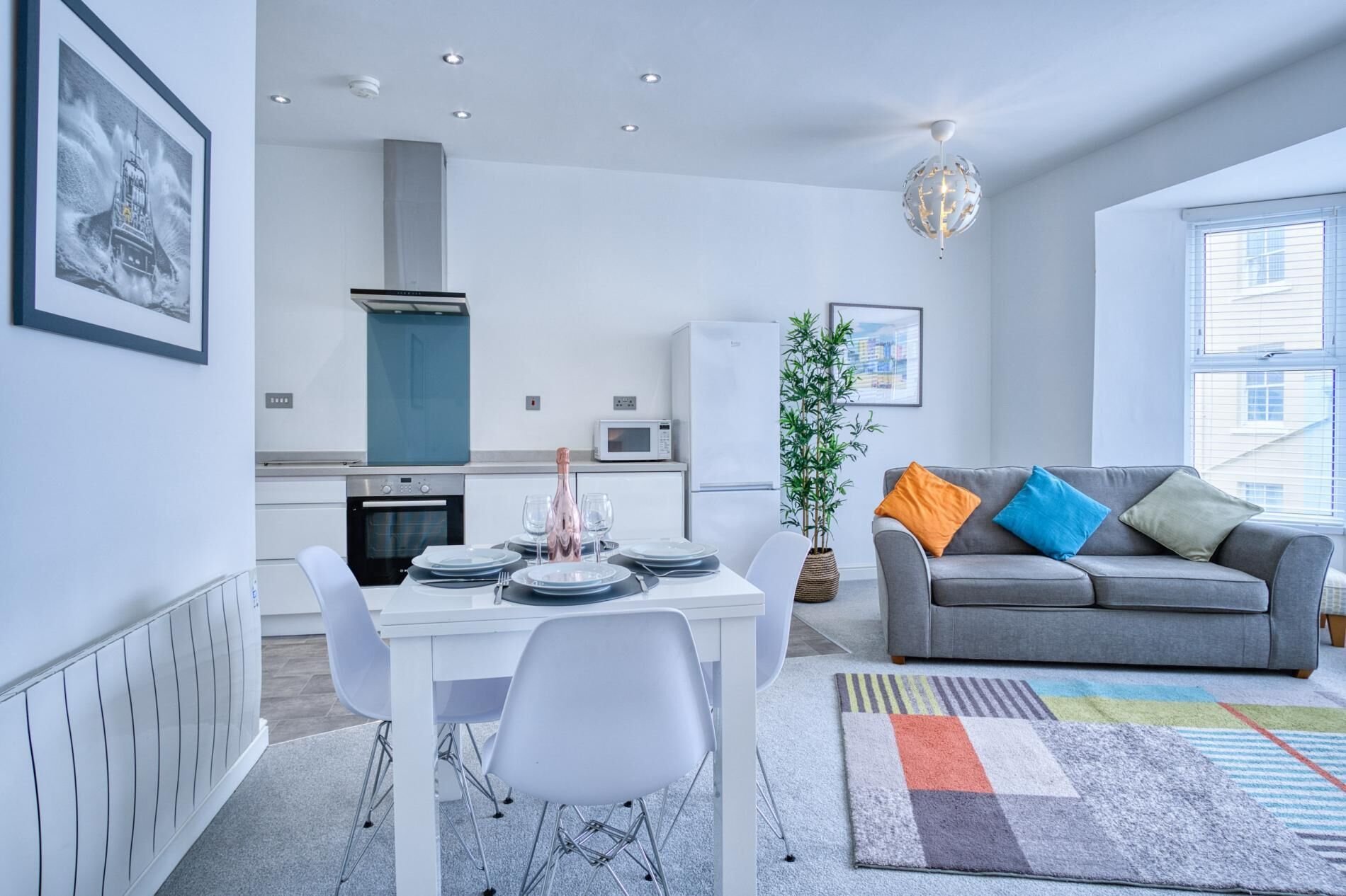 Een moderne woonkamer met een grijze bank, kleurrijke kussens, een eettafel met stoelen, een keuken met inbouwapparatuur en een groot raam.