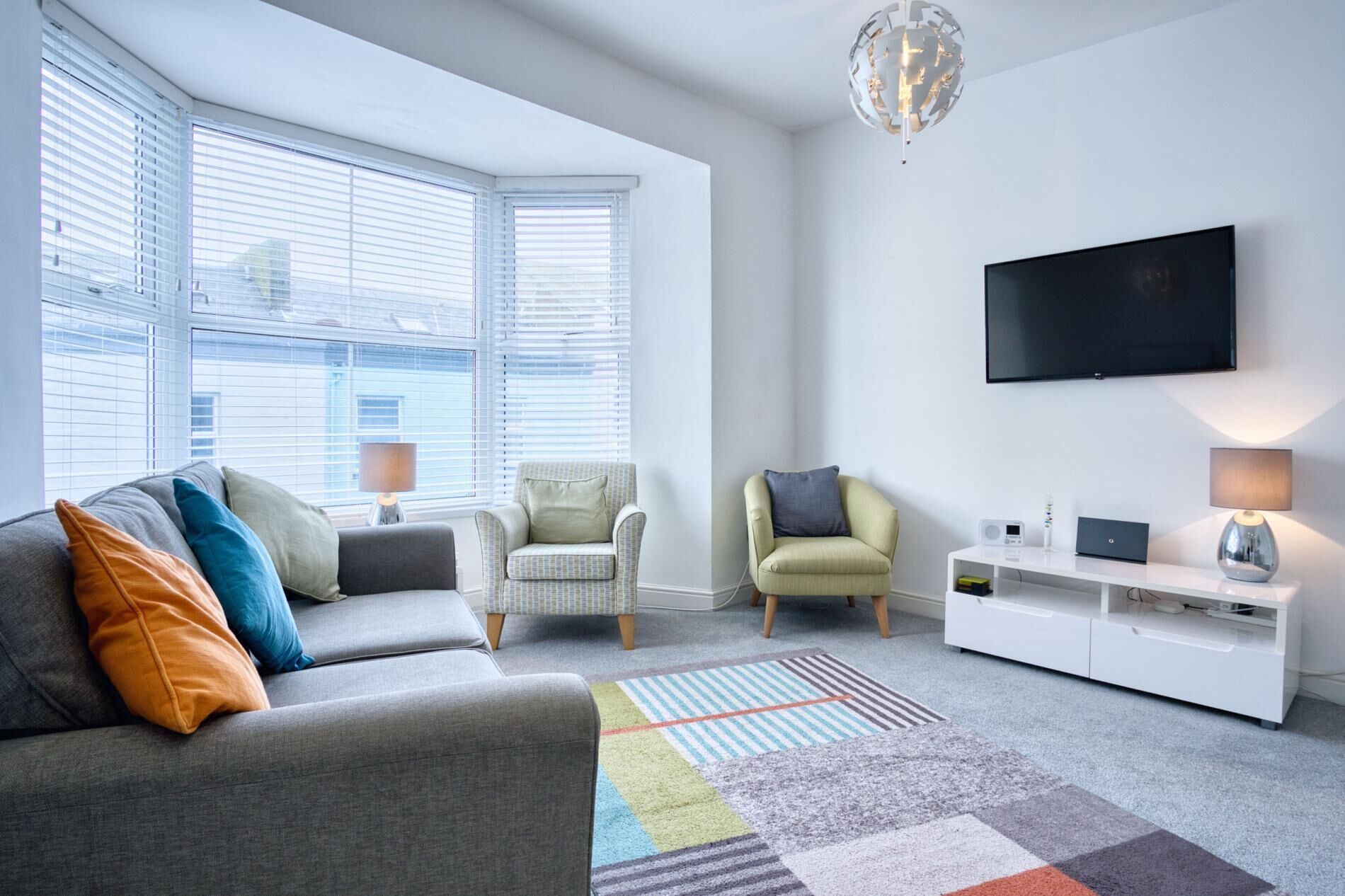 Una sala de estar moderna con un sofá gris, dos sillones, un televisor de pantalla plana, un mueble blanco para entretenimiento y una alfombra estampada.