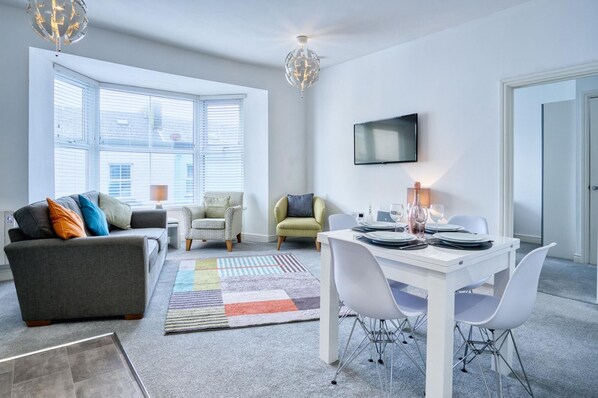 A modern living room with a grey sofa, two armchairs, a dining table set for four, and a large window with blinds. - Bay View - 2 Bedroom Apartment - Tenby (Tenby)