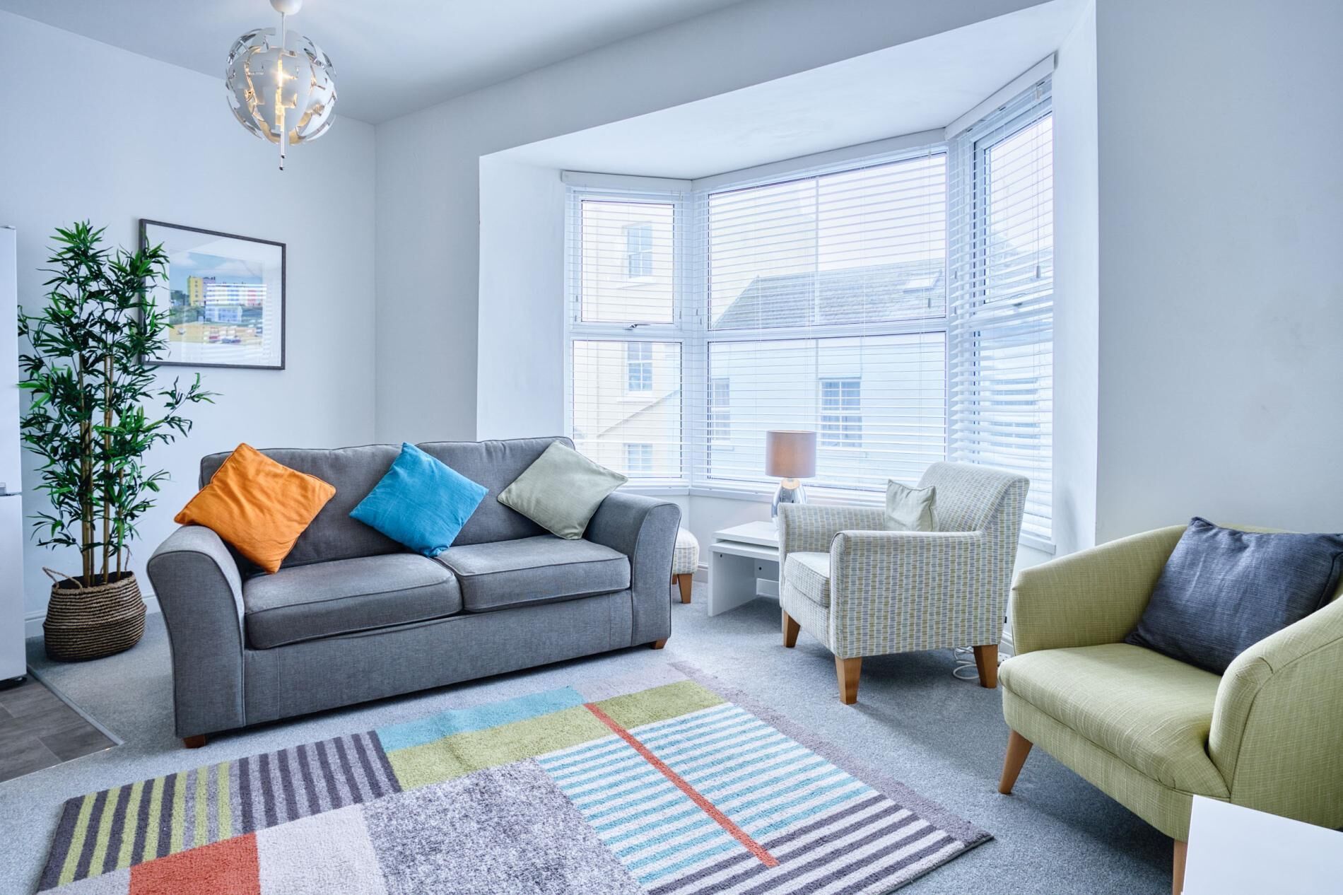 A modern living room with a grey sofa, colorful cushions, a patterned rug, and a large window with blinds.