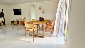 Un espacio habitable amplio con una mesa de comedor de madera y sillas anaranjadas, un televisor de pantalla plana montado en la pared y un gran ventanal con cortinas translúcidas.