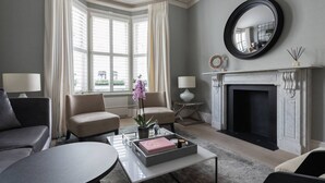Un salon moderne avec une cheminée, deux fauteuils, une table basse et une grande fenêtre avec des rideaux.