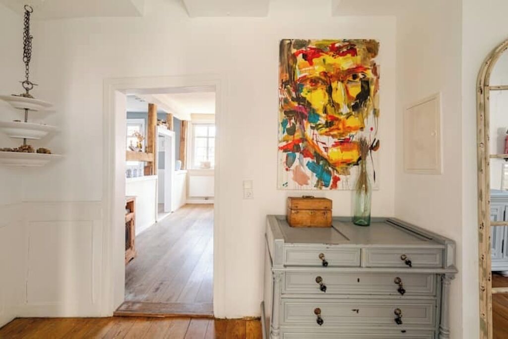 A hallway with a white wall, a gray dresser with drawers, a colorful abstract painting, and a hanging light fixture.