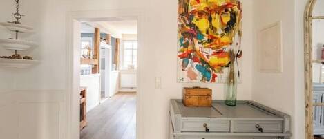 A hallway with a white wall, a gray dresser with drawers, a colorful abstract painting, and a hanging light fixture.